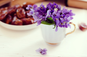 Still life with violet in white cup, old books and date fruits in a plate. Romantic floral background.