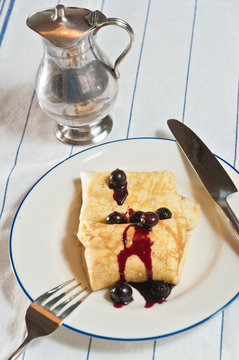 Top view, medium distance of a freshly baked, homemade blintz with lemon, blueberry sauce on a round, white plate with a blue rim and artisan knife and fork and a puter syrup container