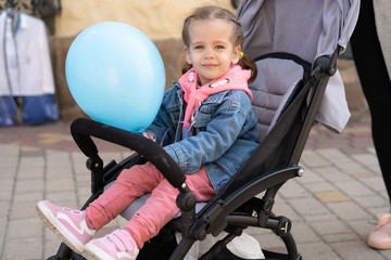 A little Caucasian girl sits in a pram and holds an inflatable ball in her hand.