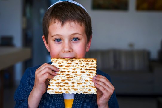 Cute Caucasian Jewish boy holding in his hands and taking a bite from a traditional Jewish matzo unleavened bread. Jewish Passover Pesach concept image.