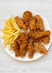 Tasty fried chicken drumsticks, spicy wings, French fries and chicken fingers on white plate over white wooden surface, top view. Flat lay, overhead, from above. Close-up.