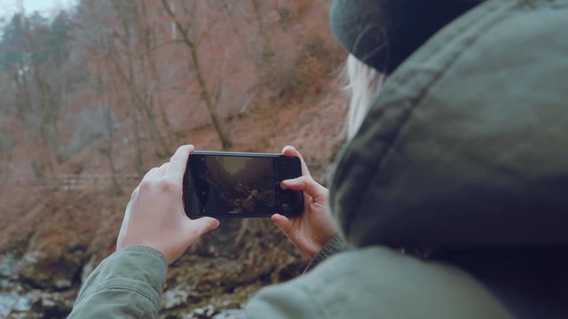 A close up of a woman taking a photo of a river running through a canyon full of rocks with her phone