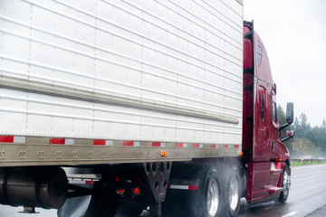 Fototapeta na wymiar Dark red big rig semi truck transporting commercial cargo in refrigerator semi trailer running on the wet highway in raining weather