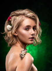 Beautiful smiling young blonde girl with flowers braided into her hair, oily body and a butterfly on her naked shoulder on a dark green gradient background. Clean, healthy skin. Close up