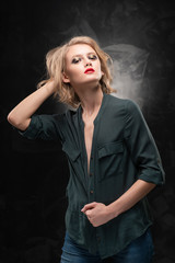 Beautiful young braless slim blonde girl with disheveled hair and aggressive makeup, wearing an unbuttoned shirt and jeans emotionally posing on a gray background. Commercial design