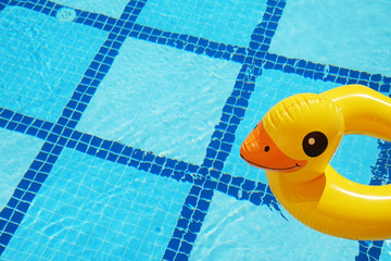Top view of inflatable duck floating in an empty swimming pool with crystal clear water and blue square tile pattern background. Close up shot of rubber ring with a lot of copy space for text.