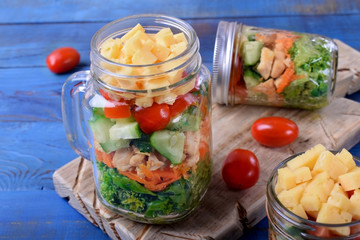 Layered salad with cheese, kale, carrot, chicken, cucumbers and cherry tomatoes in glass mason jars