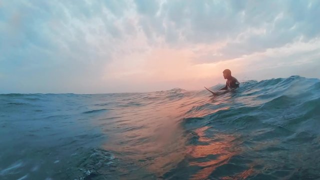 A cold water surfer is waiting in the line up in the ocean in a beautiful sunset while a wave approaches. He does a duck dive. 