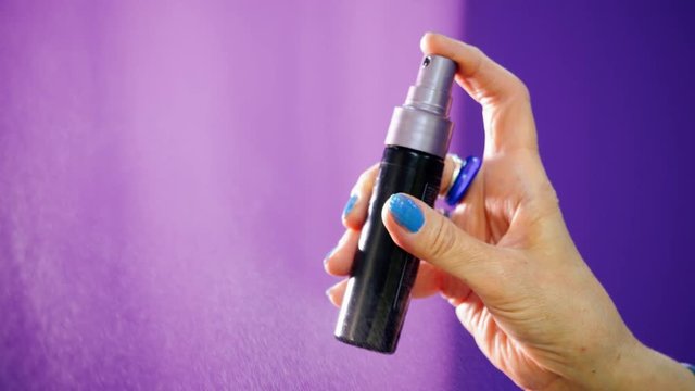 Woman's hand with spray bottle is spraying something on purple background. Close-up view female hand hold spray and push buttom. Backlit artificial light