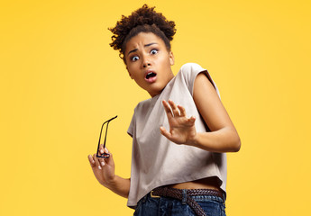 Scared girl outstretched her palm, hiding her face. Photo of african american girl wears casual outfit on yellow background. Emotions and pleasant feelings concept.