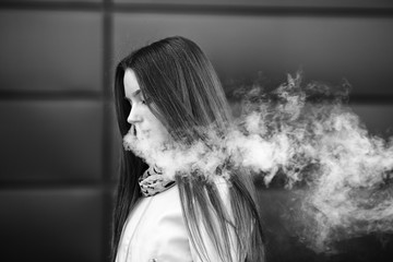 Vape teenager. Young pretty white girl in casual clothing smoking an electronic cigarette opposite modern background on the street in the spring. Bad habit. Vaping activity. Close up. Black and white.