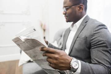 Young African American businessman in a gray suit reading a newspaper while sitting on a sofa.