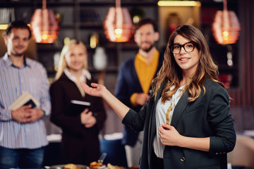 Charming businesswoman in formal wear and with eyeglasses smiling, looking at camera and showing her successful team. Restaurant interior.