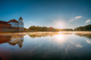 Fototapeta na wymiar Mir, Belarus. Scenic View Of Castle Complex Mir And Lake In Misty Morning Fog During Summer Sunrise. Cultural Monument, UNESCO World Heritage Site. Famous Landmark And Popular Destination
