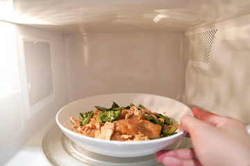 Hand man putting a white plate of stir fried noodle with pork and vegetables in microwave...