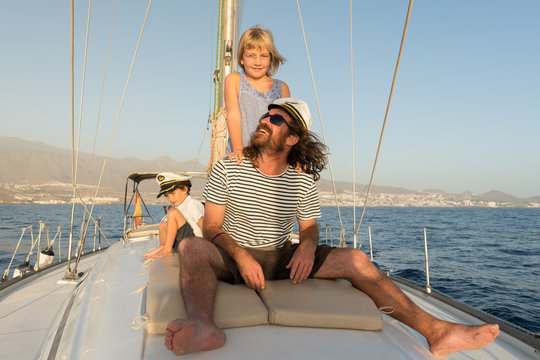 Positive father with happy kids in captain hats and sitting on deck of expensive boat floating on water in sunny day