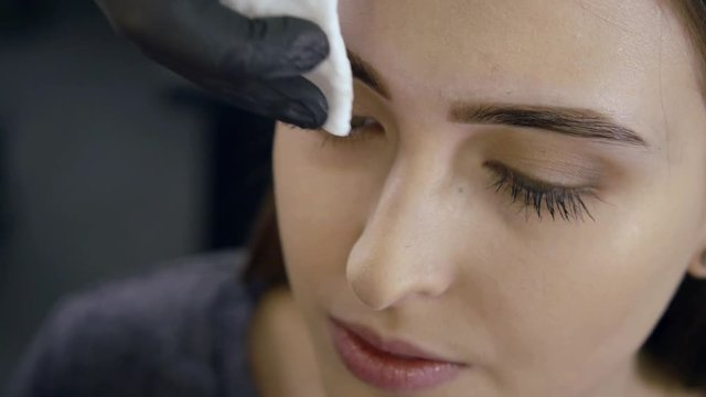 cosmetologist is wiping paint from face of female client after procedure of coloring brows