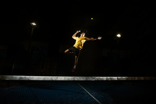 Athletic male holding racket and jumping to kick ball on tennis court in obscurity