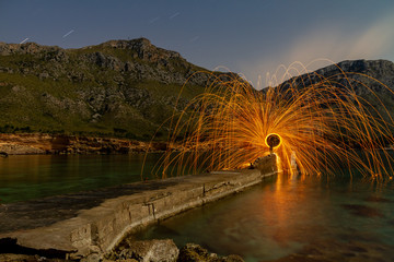 Long exposure of silhouette of human with burning round and fireworks on rocks between water near mountains in evening