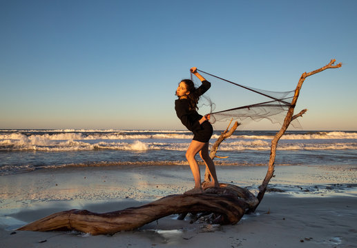Side view of young woman tangling in fishing net fixed on tree branch on sand coast with waving sea and blue sky