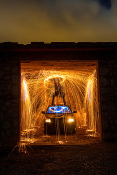 Long exposure of silhouette of human with burning circle standing on roof of automobile in shed between fireworks in obscurity
