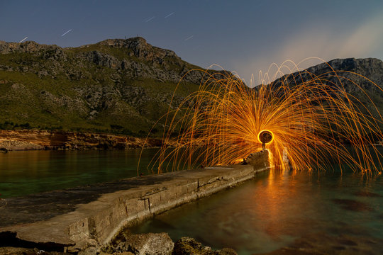 Long exposure of silhouette of human with burning round and fireworks on rocks between water near mountains in evening