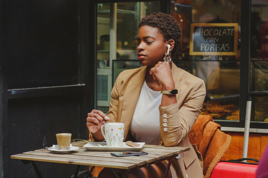 African American elegant woman with earphones looking at camera, holding mug of beverage and sitting at table in street cafe on blurred background
