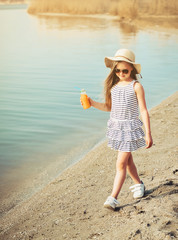 Little girl in hat walking on the beach and drinking orange juice.