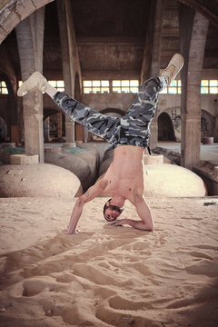 Shirtless young male performing modern dance movement on sandy ground inside aged building