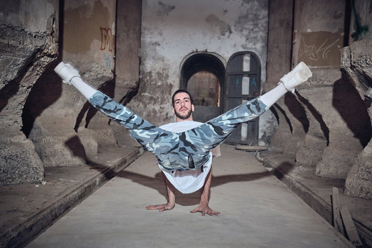 Attractive young man in stylish outfit looking at camera while performing difficult dance movement near weathered columns of aged building