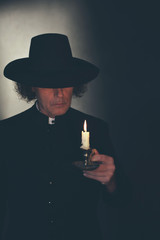 Mysterious victorian priest in black coat and hat holding candlestick.
