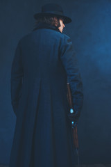 Victorian man in long black coat and hat holding rifle. Rear view.