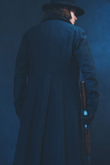 Victorian man in long black coat and hat holding rifle. Rear view.