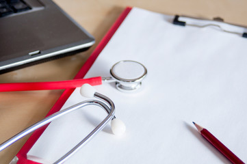 Red stethoscope and red pencil on a red clipboard on a table. Near laptop. Medical device. Treatment, health care. Heart examination. Studying the pulse.