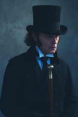 Edwardian man in long black coat and hat holding cane.