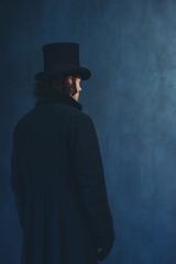 Edwardian man in long black coat and hat standing towards grey wall.