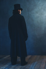 Edwardian man in long black coat and hat standing towards grey wall.