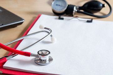 Red stethoscope on a red clipboard. Near laptop and tonometer. Copy space. Medical device. Treatment, health care. Heart examination. Studying the pulse. Close-up.