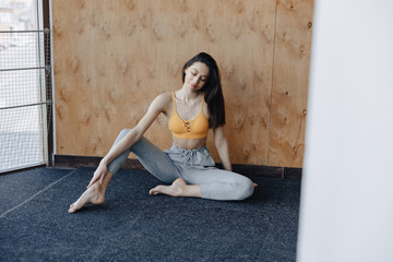 Young attractive fitness girl sitting on the floor near the window on the background of a wooden wall, resting on yoga classes