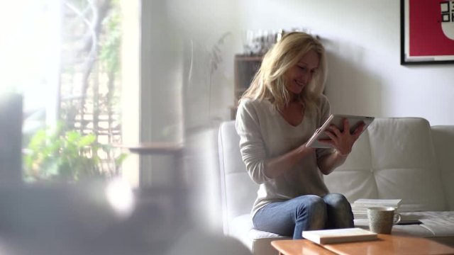 Medium shot of a smiling mature woman drinking coffee and using a digital tablet while sitting on the sofa at home