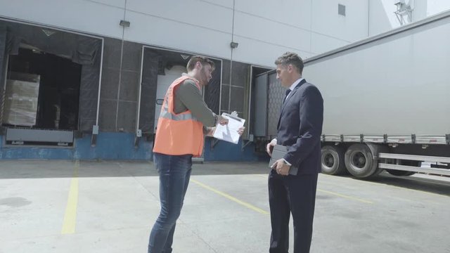 Medium shot of warehouse worker and businessman discussing over a clipboard near lorry