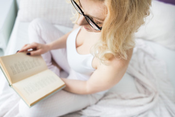 Attractive blonde woman in glasses with black rim sits on the edge of the bed at home with a book and reads