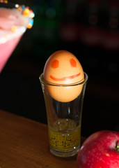 brown egg with painted smile and eyes in glass near red apple on wooden table in bar