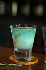 side view of shot glass with blue alcohol drink on dried orange slice with rosemary on wooden table in bar