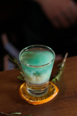 high angle view of shot glass with blue alcohol drink on dried orange slice with rosemary on wooden table in bar