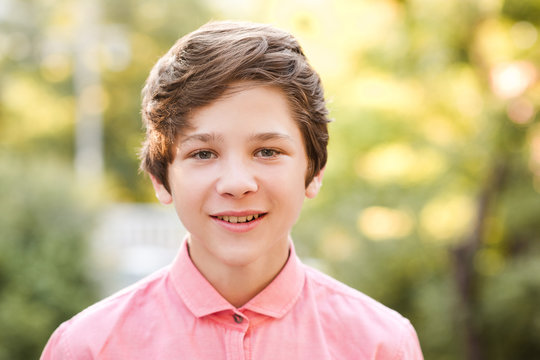 Smiling Blond Boy 15-16 Year Old Stdning In Park Over Nature Green Background. Wearing Pink Shirt. Looking At Camera. Summer Season.