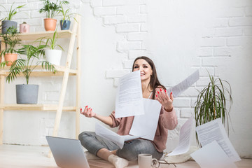 A young happy woman throws up a bunch of pieces of paper and documents sitting near the laptop, the freelance girl is happy and in a good mood. excitement and inspiration - obrazy, fototapety, plakaty