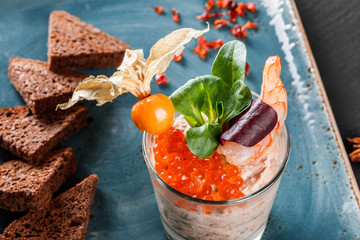 Snack from shrimps and red caviar with black bread, decorated with physalis and greens on plate over black background. Healthy food
