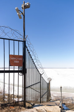 Guarded zana with video surveillance. Barbed wire used on top of a metal fence, with a sign in Russian - Forbidden Zone - hanging on the side. Winter concept. The reservoir is covered with ice.