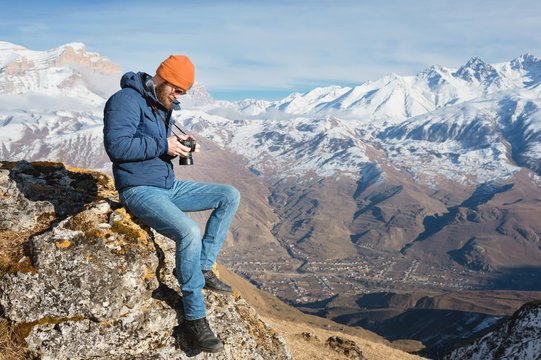 Portrait bearded male photographer in sunglasses and warm jacket with reflex camera in his hands looks at the photos on the camera while sitting on stone in the background of snow-capped mountains